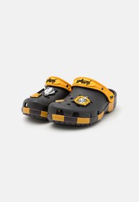 Svarta crocs med gula remmar, med dekorativa lappar: en grävling och ett vapen. Texten "hufflepuff" på remmen; texturerad yta.
