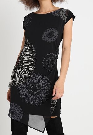 Kobieta w czarnej sukience z dużymi, misternymi szarymi wzorami kwiatowymi i asymetrycznym dołem, w połączeniu z czarnymi, wysokimi botkami za kolano.