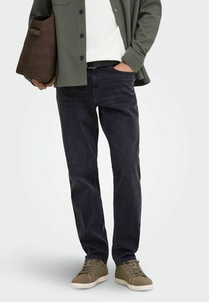 Mand iført mørke straight-leg jeans, olivengrønne sneakers, grøn skjorte over en hvid t-shirt, holder en brun taske med den ene hånd i lommen.