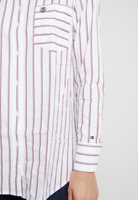 Chemise blanche à boutons avec des rayures verticales rouges et bleu marine, comportant une poche poitrine et un logo brodé sur la poche. Le poignet a un détail rayé.