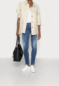 Chaqueta utility beige con botones en la parte delantera y bolsillos, blouse blanca, jeans azules ajustados y bolso negro combinados con zapatillas blancas.