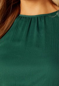 Top plisado verde con cuello alto, que presenta una superficie texturizada y una tela suave y fluida. Diseño casual, ideal para estilos en capas.