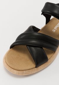 Sandale en cuir noir avec des sangles croisées, semelle texturée et un coussin de pied de type liège brun clair. La partie supérieure présente une finition lisse et des coutures discrètes.