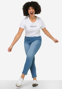 Camiseta blanca con el texto "Jeggings", combinada con jeggings azules. La modelo lleva zapatillas blancas, sonríe y tiene el cabello rizado, en un fondo sencillo.
