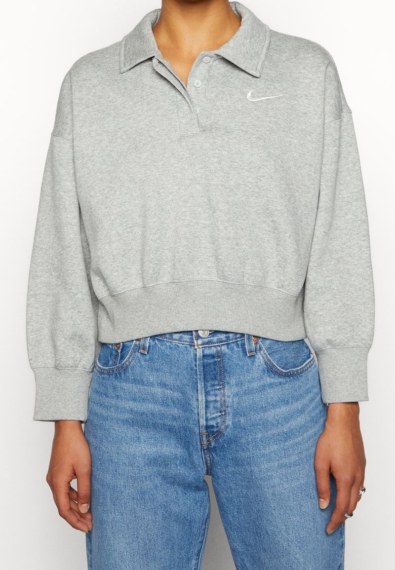 Grå kortärmad polo sweatshirt med ribbad nederkant och ärmslut, med en knappslå och en liten vit Nike-logotyp på bröstet.