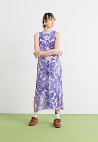 Dlouhé šaty bez rukávů z jasně fialové tkaniny s květinovými vzory, bočními rozparky a mírně průhlednou texturou. Nosené s hnědými botami.