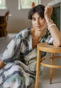 Žena s vlnitými tmavými vlasmi a náušnicami, oblečená v zelených a sivých vzorovaných šatách, opierajúca si ruku o drevenú stoličku v interiéri.