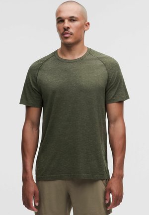 Uomo con testa rasata che indossa una camicia a maniche corte verde oliva texturizzata e pantaloni marrone chiaro, in piedi davanti a uno sfondo semplice.
