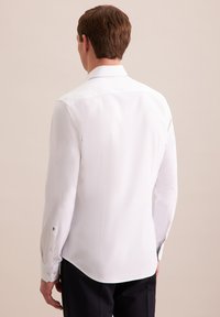 Osoba z krótkimi brązowymi włosami stoi tyłem, ubrana w białą koszulę o dopasowanym kroju, na jasnym beżowym tle.