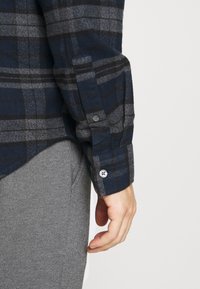 Camicia in flanella a quadri blu navy e grigia con polsini abbottonati, tessuto testurizzato e orlo curvo, abbinata a pantaloni di maglia grigi.