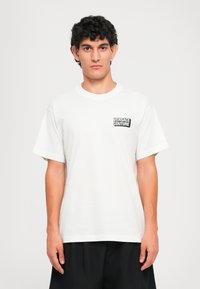 Homem com cabelo encaracolado escuro veste uma t-shirt branca da Versace Couture e calças pretas, posando contra um fundo claro e simples.