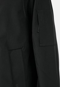 Giacca nera impermeabile con tessuto dalla texture liscia, tasca sul petto e logo sulla manica. Il design è minimalista e funzionale.