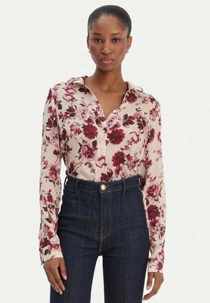 Donna con una camicetta bianca a stampa floreale rosso scuro e jeans blu scuro a vita alta, in piedi davanti a uno sfondo chiaro uniforme.