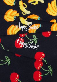 Chaussettes marines avec des motifs colorés de bananes et de cerises, portant le logo "Happy Socks" en blanc. Texture lisse, design en tissu tricoté.