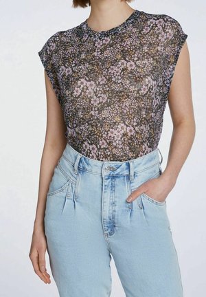 Frau trägt ein transparentes schwarzes Blumentop ohne Ärmel, in eine hoch taillierte hellblaue Jeans gesteckt, mit einer Hand in der Tasche.