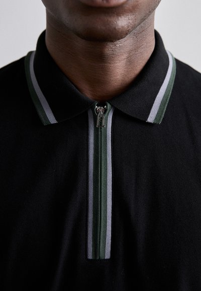 Juodas polo marškinėliai su užtrauktuku, kuriuose yra žalios ir pilkos juostelės akcentai ant apykaklės ir užtrauktuko. Pagaminti iš minkštos medžiagos, minimalus tekstūros.