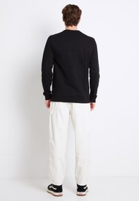 Uomo che indossa una maglietta nera a maniche lunghe, pantaloni bianchi e sneakers nere, in piedi di fronte a un muro bianco.