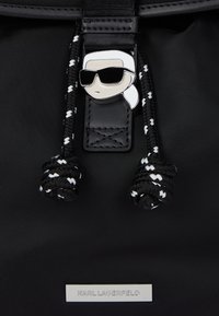 Μαύρο σακίδιο πλάτης με πλεγμένες κορδόνια και μέταλλο έμβλημα με πρόσωπο που φορά γυαλιά ηλίου και λευκό μαντήλι, επωνυμίας "Karl Lagerfeld."
