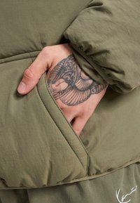 Olivgrüner, gepolsterter Mantel mit einer Seitentasche, aus strukturiertem Stoff und mit elastischen Bündchen. Eine Hand mit einem detaillierten Tattoo ist sichtbar.