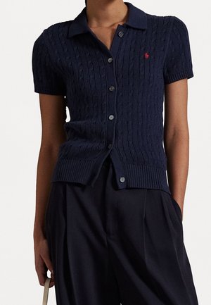 Cardigan bleu marine à manches courtes en maille torsadée avec des boutons, un col et un logo rouge brodé, associé à un pantalon noir à jambes larges.