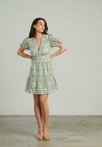 Vestido de encaje en verde menta con escote en V profundo, mangas abullonadas y falda en capas. Presenta patrones florales y un cuerpo ajustado.