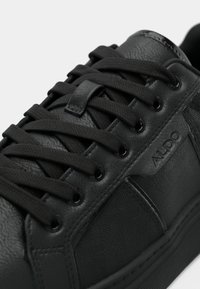 Črni tekaški copat iz sintetičnega usnja s ploskimi črnimi vezalkami, teksturiranimi stranskimi ploščami in majhnim vtisnjenim logotipom. Gladek površinski material z nevsiljivimi vzorčnimi poudarki.