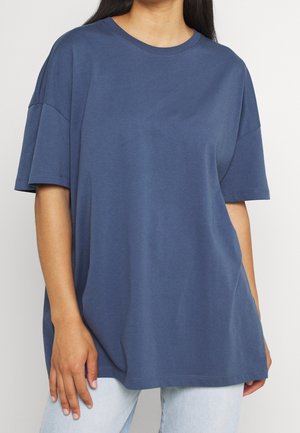 Henkilö, joka käyttää löysää, lyhythihaista sinistä t-paitaa ja vaaleanpesuista farkkuja, seisoo yksinkertaista vaaleaa taustaa vasten.