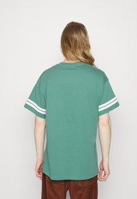 Person med långt hår som bär en grön kortärmad skjorta med vita dubbla ränder på ärmarna, vänd bort från kameran mot en helt vit bakgrund.