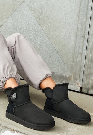 Osoba w jasnoszarych spodniach dresowych i czarnych botkach z ozdobnymi guziczkami, siedząca na betonowej podłodze przy metalowej ścianie.