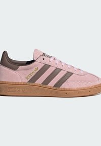 Sapatilhas de camurça cor-de-rosa com detalhes em pele castanha, três riscas castanhas, atacadores brancos e uma sola de borracha texturizada. Apresenta um detalhe de logotipo dourado.