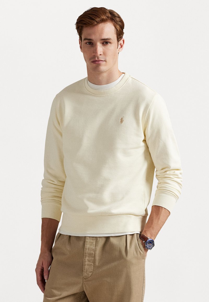 Homem com um suéter de malha creme por cima de uma t-shirt branca, calças bege e um relógio de pulso com um mostrador azul e uma bracelete de couro castanho.