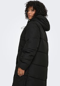 Musta toppatakki hupulla, jossa tikattu pinta, vetoketju- ja niittikiinnitykset, suuret sivutaskut ja nyörikiristysaukko kauluksessa.