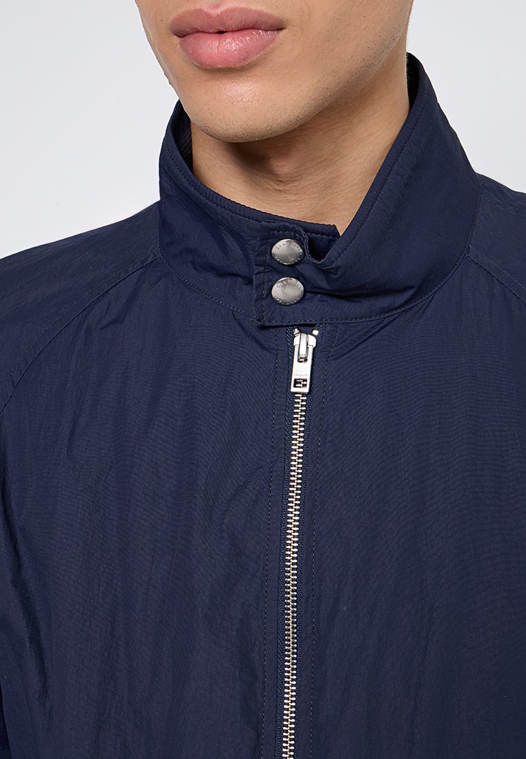 Dunkelblaue Jacke mit hohem Kragen, silbernen Druckknöpfen und einem zentralen Reißverschluss. Hergestellt aus leichtem Stoff mit strukturierter Oberfläche.