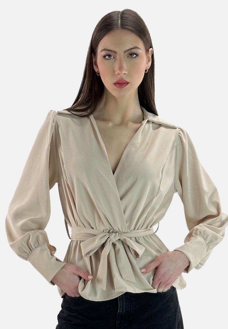Blusa avvolgente beige con maniche lunghe a sbuffo, dettaglio con cintura da annodare e tessuto liscio e lucido. Indossata con jeans scuri, in piedi contro uno sfondo semplice.