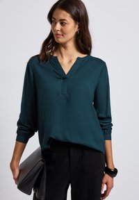 Blusa verde acqua realizzata in tessuto morbido, con scollatura a V, maniche arricciate e vestibilità comoda. Indossata con pantaloni neri e una pochette nera.