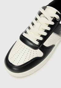 Zwarte en crème sneaker met geperforeerde details, leren panelen met textuur, witte veters en een platte rubberen zool.