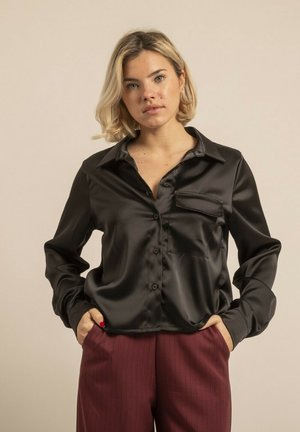 Blouse en satin noir avec un col, une fermeture par boutons et une poche poitrine. Manches longues avec une texture lisse et brillante.
