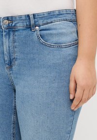 Ljusblå denimjeans med små spridda rhinestones, framfickor, bältesöljer, knappstängning och en synlig hand som vilar vid låret.