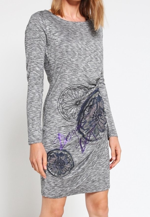 Femme portant une robe grise à manches longues avec des motifs floraux noirs et des motifs circulaires, ainsi qu'une écriture cursive violette sur le devant.
