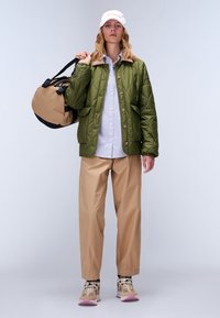 Giacca verde trapuntata con colletto marrone, camicia a righe blu, pantaloni beige, con una borsa a tracolla beige e nera, e indossando sneaker beige.