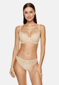 Beżowy stanik koronkowy z fiszbinami i regulowanymi ramiączkami, w komplecie z dopasowanym przezroczystym thongiem z kwiatowymi wzorami i małym detalem w postaci kokardki.