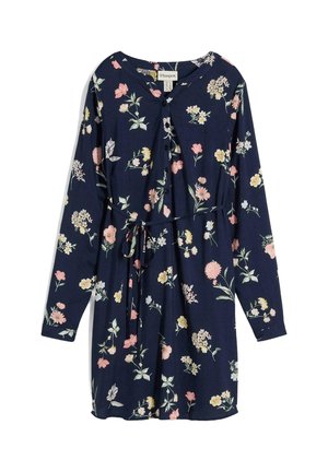 Femme portant une tunique bleu marine à imprimé floral multicolore, les manches retroussées et un legging noir, debout avec un bras passé devant la taille.