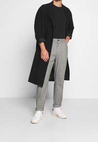 Abrigo negro con mangas enrolladas, camiseta negra ajustada debajo y pantalones de cuadros grises. Zapatillas blancas con ligeros acentos.