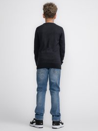 Jersey de punto oscuro, jeans de mezclilla azules, zapatillas deportivas negras, visto desde atrás contra un fondo liso. Cabello corto y rizado visible.