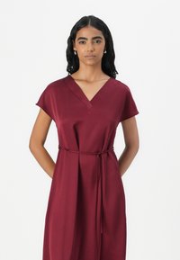 Robe bordeaux en tissu lisse avec un décolleté en V, des manches courtes et un lien à la taille, mettant en avant une silhouette détendue.