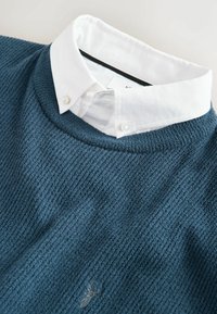 Gestrickter Pullover in dunklem Petrol mit gerippter Textur, darunter ein weißes Hemd mit Kragen. Dezentes Logo-Detail nahe dem Saum.