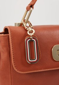 Sac en cuir marron texturé avec quincaillerie dorée et un porte-clé rectangulaire comportant des rayures en émail rouge, blanc et noir.