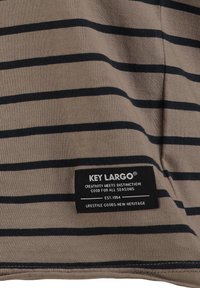 Tejido marrón claro con rayas horizontales negras; presenta una etiqueta de marca con texto sobre un fondo negro. La textura parece suave y informal.