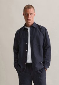 Marineblauw knoopshirt van een gestructureerde stof, met lange mouwen, één borstzak en witte knopen. Draagt over een eenvoudig wit shirt.