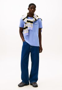 Jasnoniebieski t-shirt z krótkim rękawem z grafiką na piersi, zestawiony z luźnymi, ciemnoniebieskimi dżinsami. Na ramionach przewieszony pasiasty sweter z dzianiny.